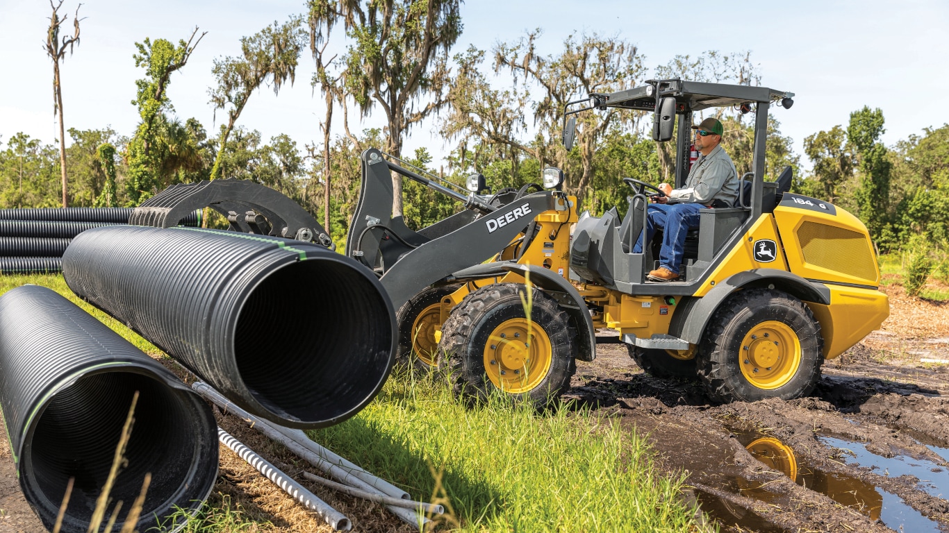 Chargeuse à roues compacte John Deere 184G soulevant un tuyau noir.